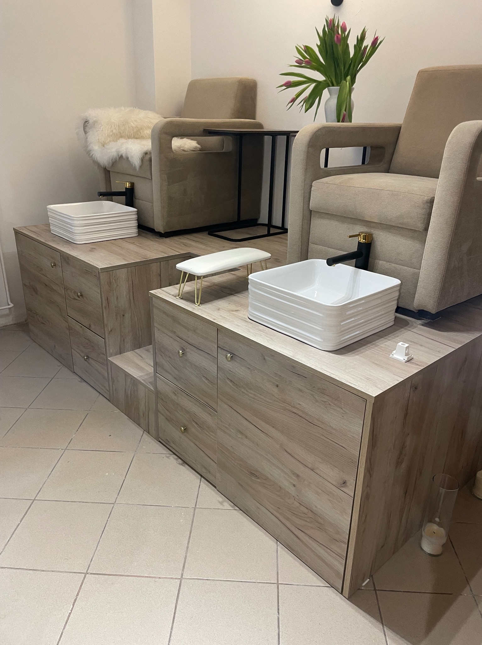 Nowoczesne stanowiska do pedicure z drewnianymi szafkami, białymi misami i beżowymi fotelami. Wazon z tulipanami dodaje elegancji. Jasne płytki podłogowe.