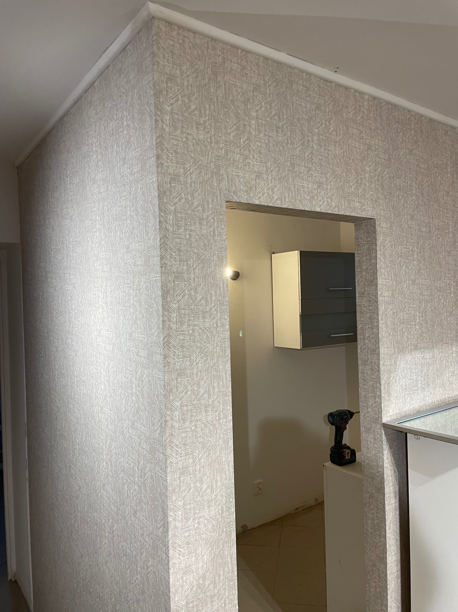 Ściana wykończona tapetą w odcieniach szarości z wzorem, widoczny fragment sufitu z listwą. W tle szafka i wkrętarka na blacie. Ujęcie z bliska.