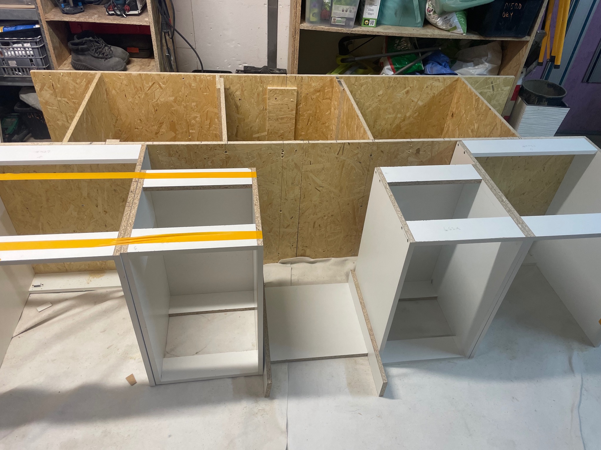 Stolarz w trakcie pracy: białe szafki z płyty meblowej, częściowo zmontowane, na tle regału z płyty OSB. Widoczne detale montażowe i żółta taśma zabezpieczająca.