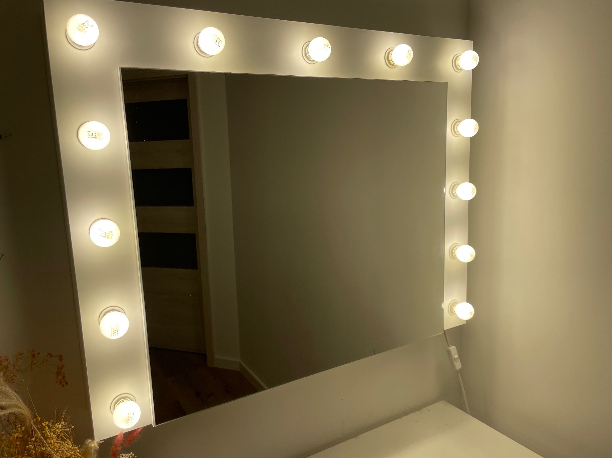 Białe lustro do makijażu z żarówkami LED, odbijające fragment pokoju z drewnianą podłogą. Widoczny włącznik i kabel zasilający. Ciepłe światło żarówek.