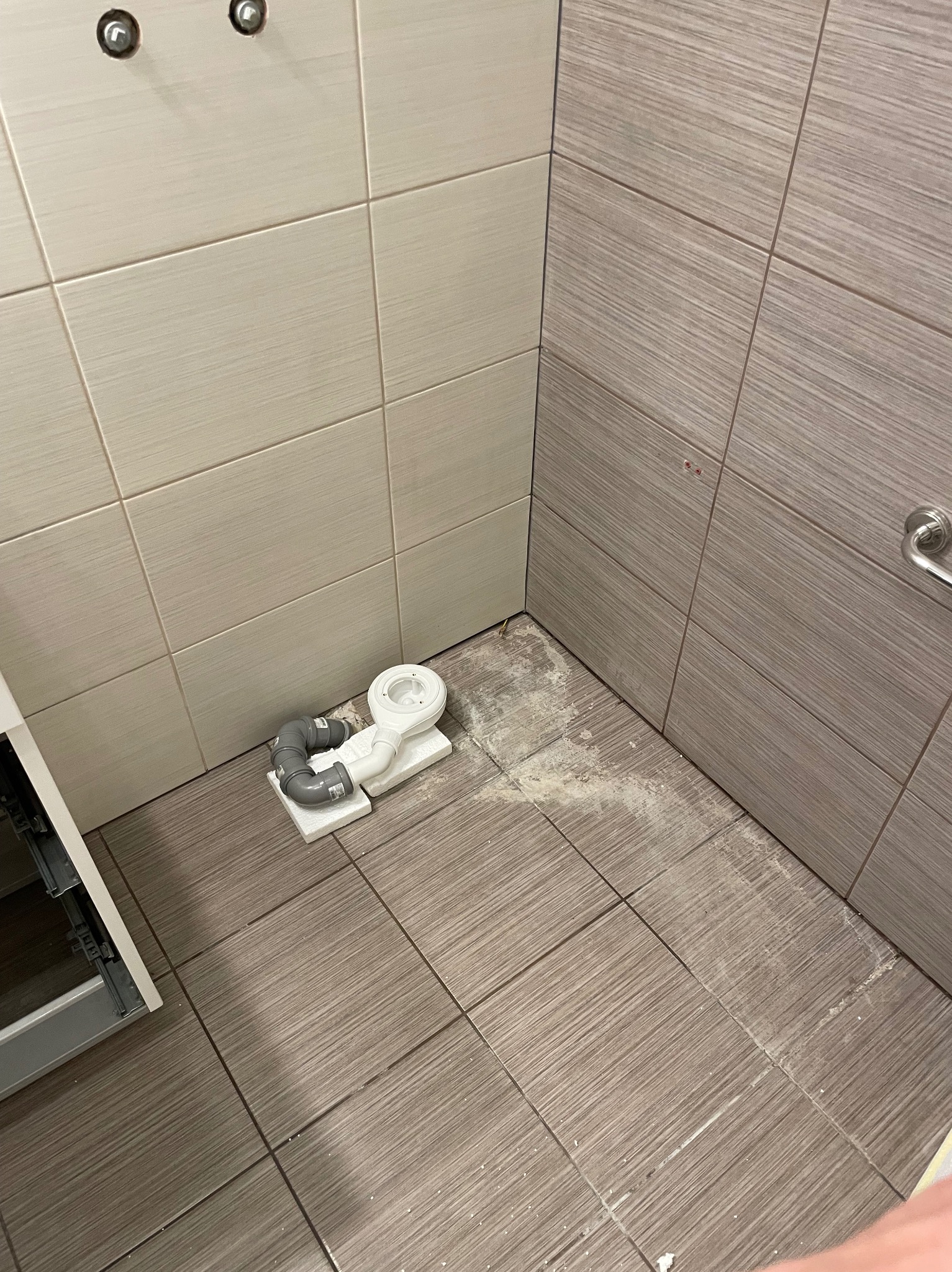 Ukończona instalacja odpływu liniowego w łazience z szarymi płytkami. Widoczne elementy syfonu i fragment szafki pod umywalkę. Przygotowanie do dalszych prac.
