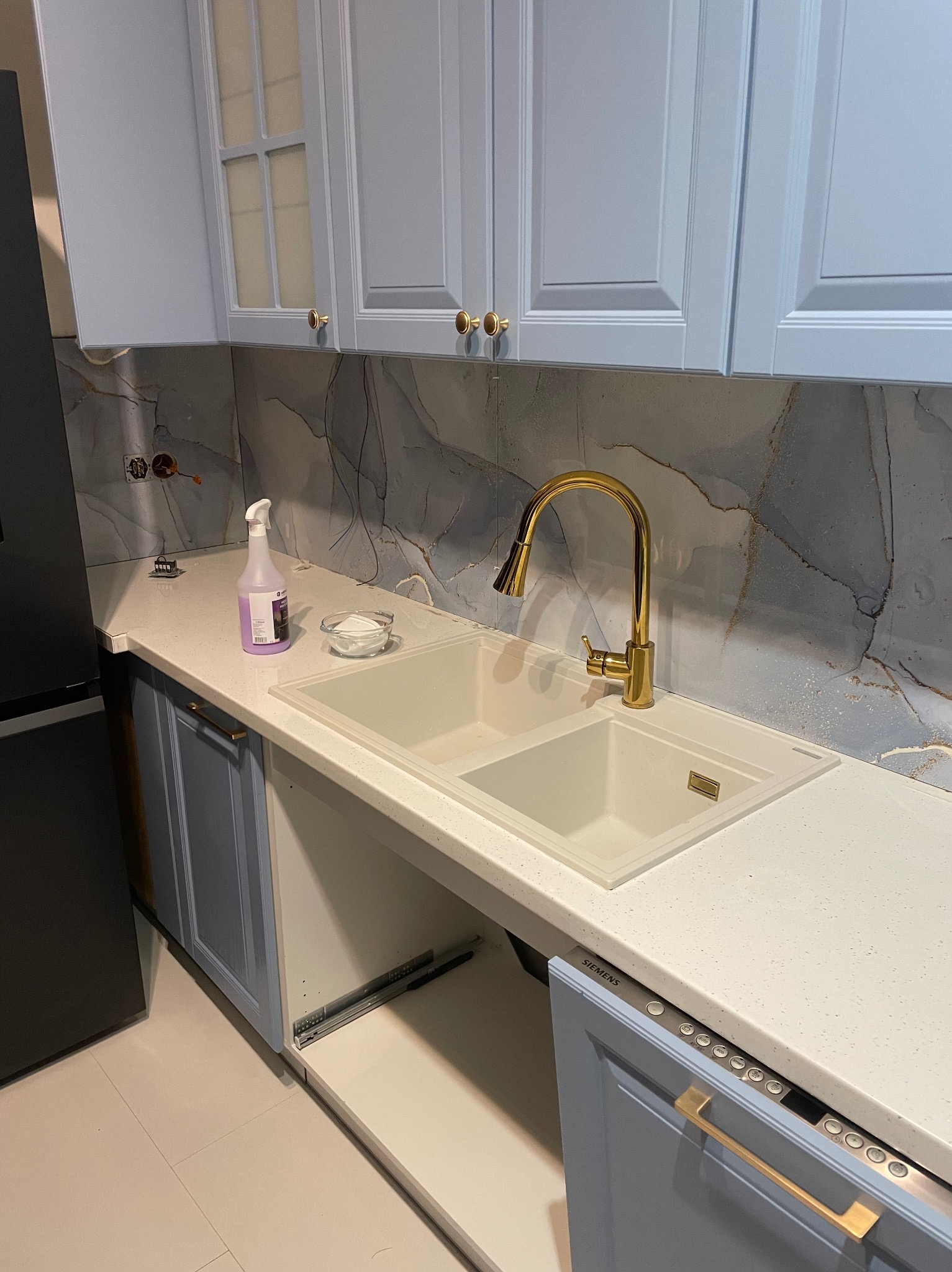 Błękitna zabudowa kuchenna z marmurowym blatem i złotą armaturą. Widoczny fragment lodówki, szafki z przeszkleniami i sprzęt AGD. Nowoczesny design.