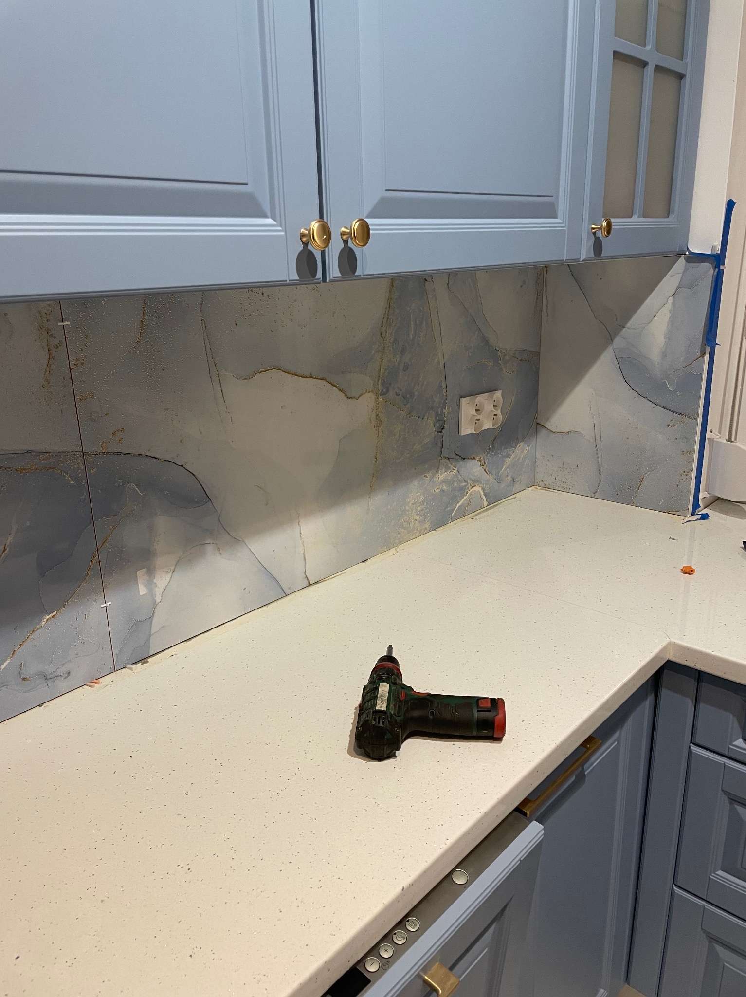 Niebieska zabudowa kuchenna z marmurowym wzorem na ścianie. Na blacie leży wiertarka. Widoczne złote uchwyty szafek i gniazdko elektryczne.