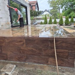 DarBruk Brukarstwo - Olszewnica Stara - Mycie wodą podestu z brązowej kostki, z widoczną fakturą drewna, na tle domu z dużymi oknami i zieleni. Prace brukarskie w toku, z użyciem węża i łopaty.