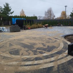 DarBruk Brukarstwo - Olszewnica Stara - Układanie wzorzystej kostki brukowej na posesji. Widoczny fragment okręgu z piaskiem, paleta z kostką i pracownik w trakcie pracy. Tło: ogrodzenie i zieleń.