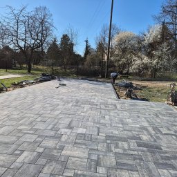 DarBruk Brukarstwo - Olszewnica Stara - Ułożona szara kostka brukowa na posesji, widoczny pracownik i sprzęt budowlany, w tle drzewa i błękitne niebo. Nietypowy wzór ułożenia kostki.