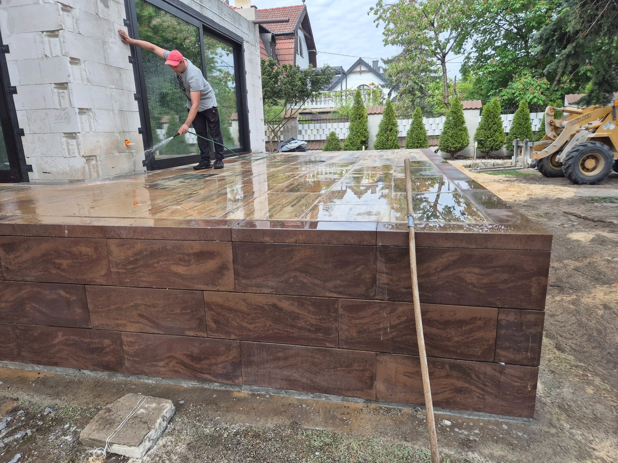 Mycie wodą podestu z brązowej kostki, z widoczną fakturą drewna, na tle domu z dużymi oknami i zieleni. Prace brukarskie w toku, z użyciem węża i łopaty.
