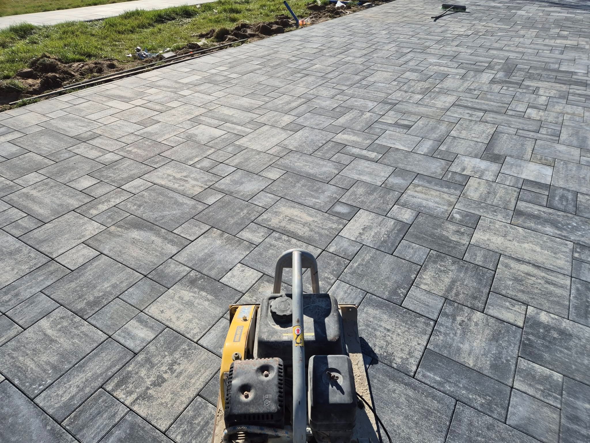 Ułożona kostka brukowa w odcieniach szarości, z widoczną zagęszczarką w dolnej części kadru. W tle trawnik i świeżo przygotowany teren pod dalsze prace.