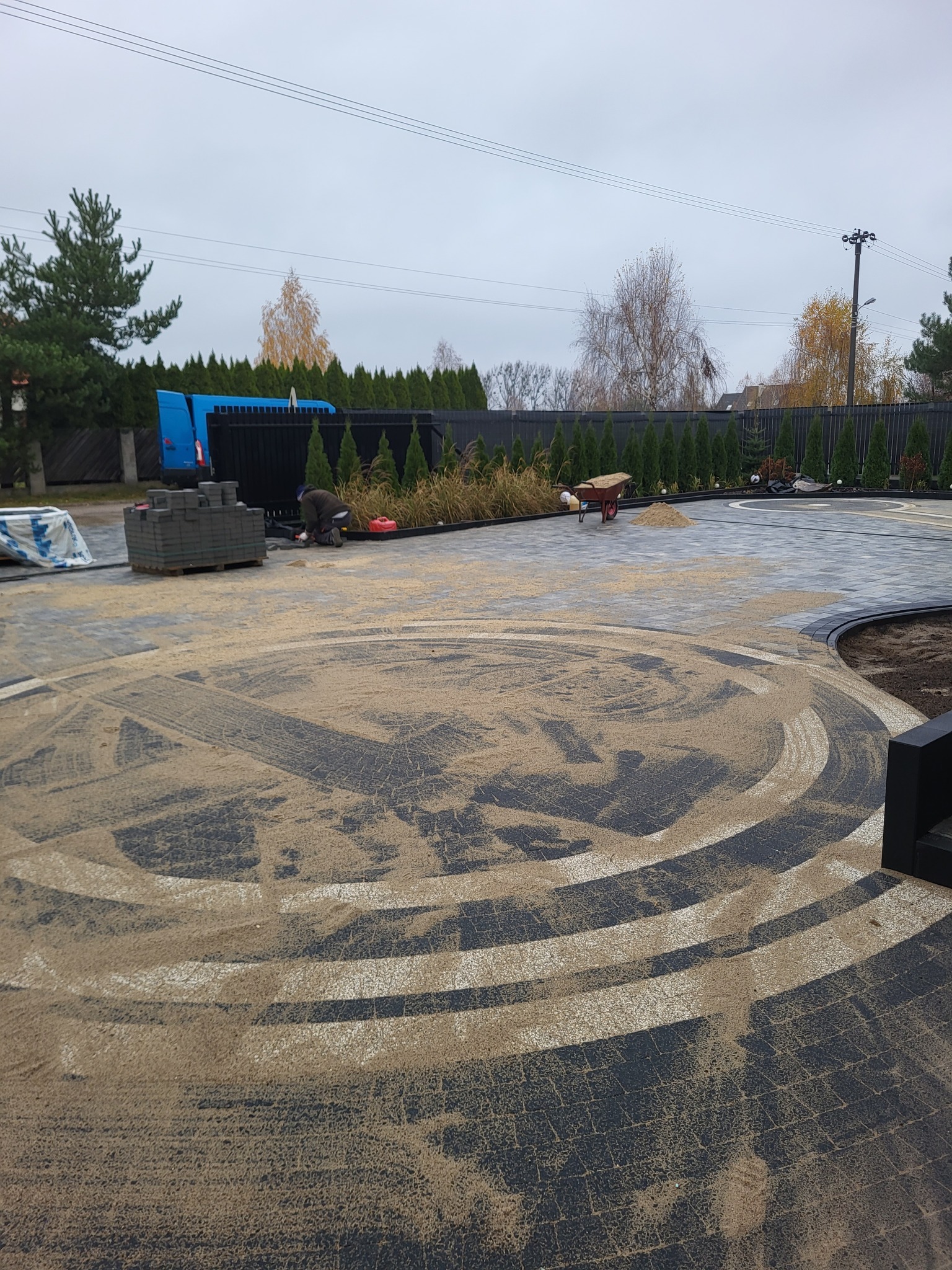 Układanie wzorzystej kostki brukowej na posesji. Widoczny fragment okręgu z piaskiem, paleta z kostką i pracownik w trakcie pracy. Tło: ogrodzenie i zieleń.