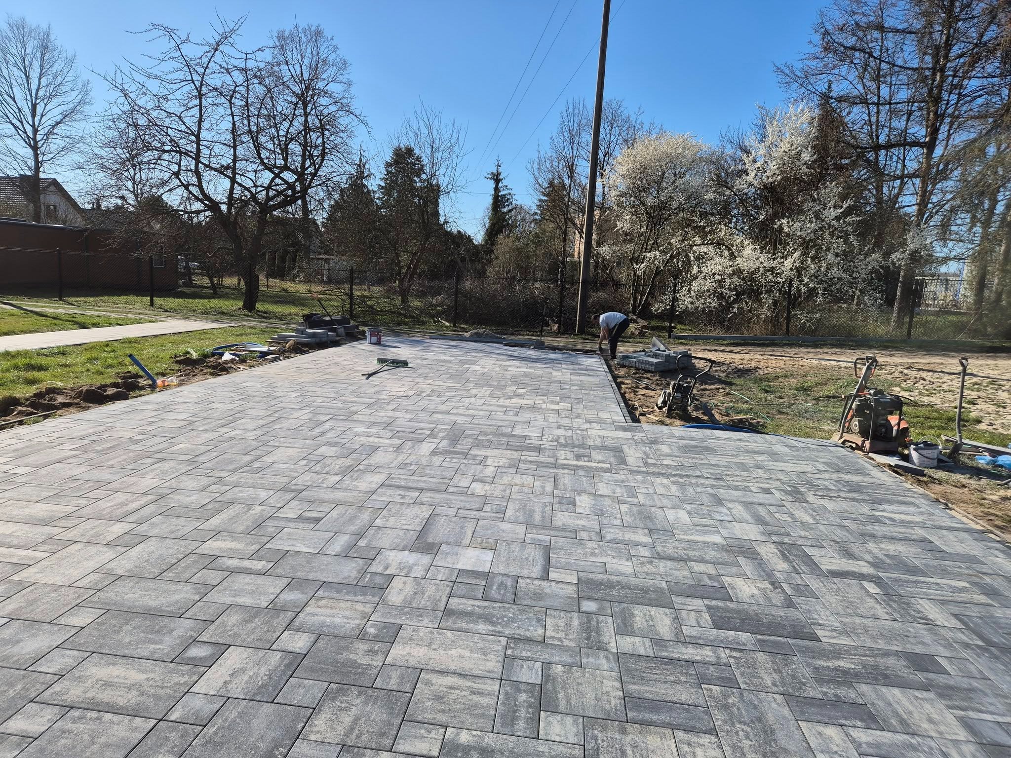 Ułożona szara kostka brukowa na posesji, widoczny pracownik i sprzęt budowlany, w tle drzewa i błękitne niebo. Nietypowy wzór ułożenia kostki.