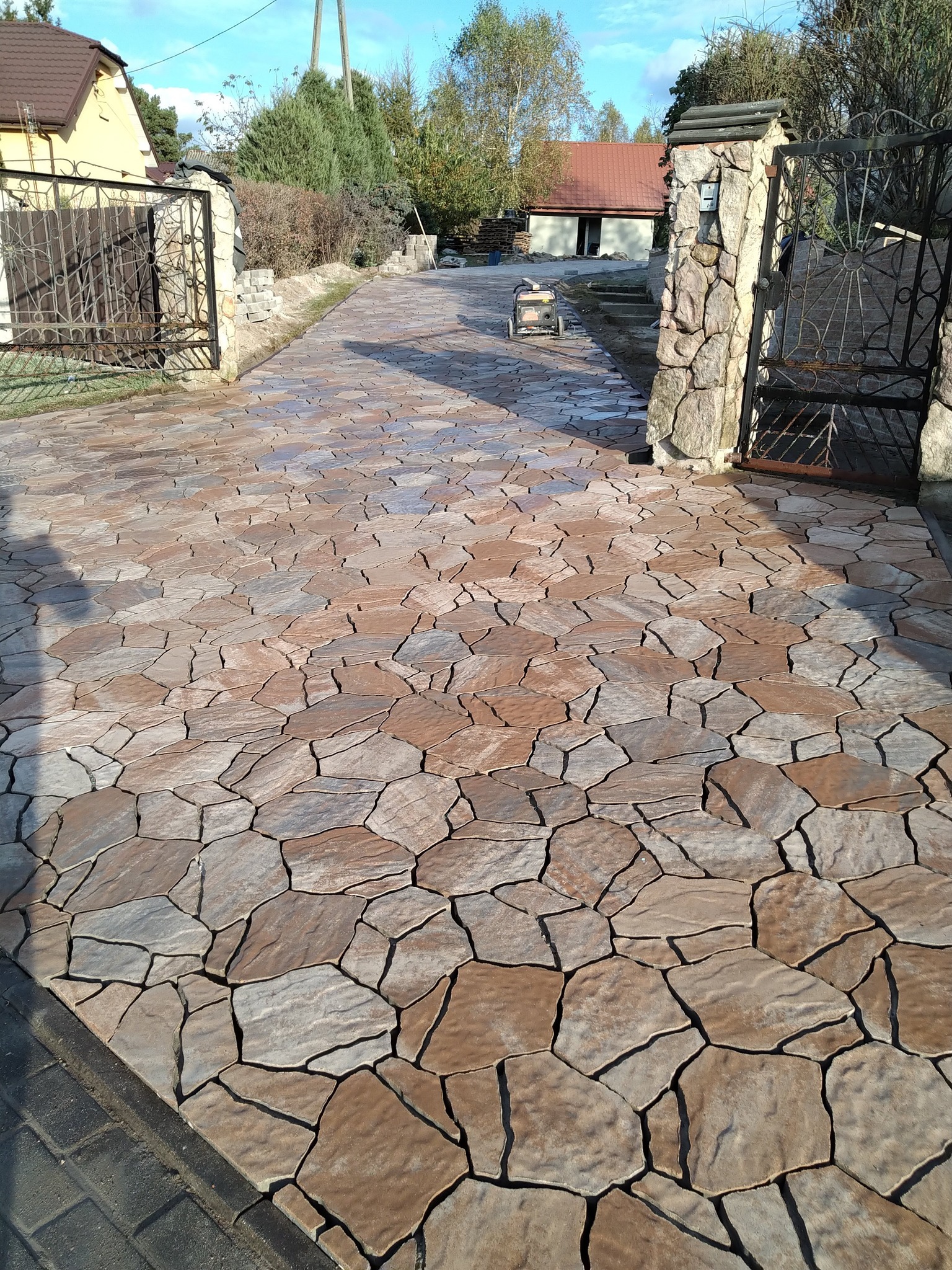 Ułożona kostka brukowa o nieregularnym kształcie, prowadząca od bramy wjazdowej do domu. Widoczne kamienne słupki bramy i fragment domu w tle.