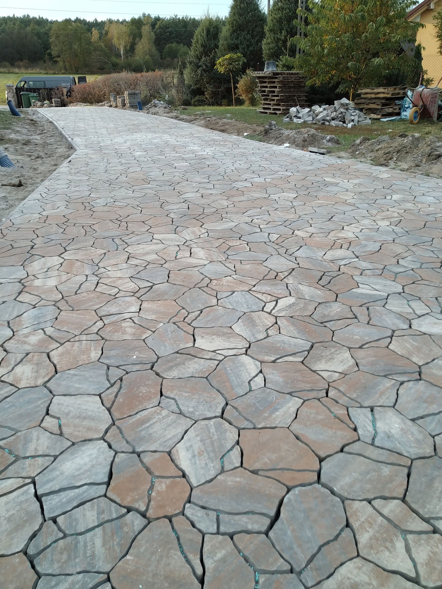 Ścieżka z nieregularnej kostki brukowej w odcieniach szarości i beżu, prowadząca przez posesję. W tle widoczne drzewa i materiały budowlane.