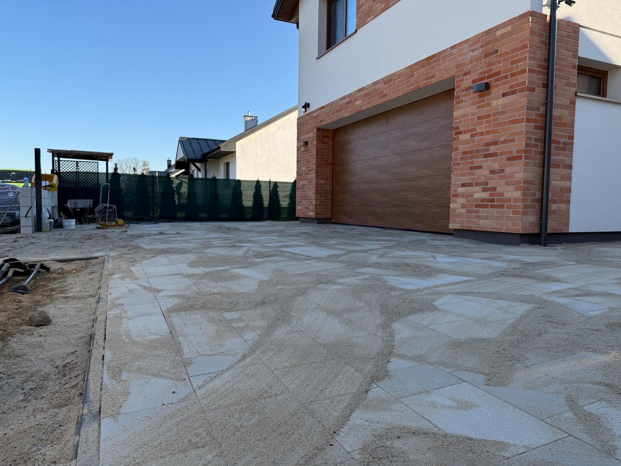Jasnoszara kostka brukowa na podjeździe domu jednorodzinnego, częściowo zasypana piaskiem. W tle widoczny budynek z brązową bramą garażową i ceglanym wykończeniem.