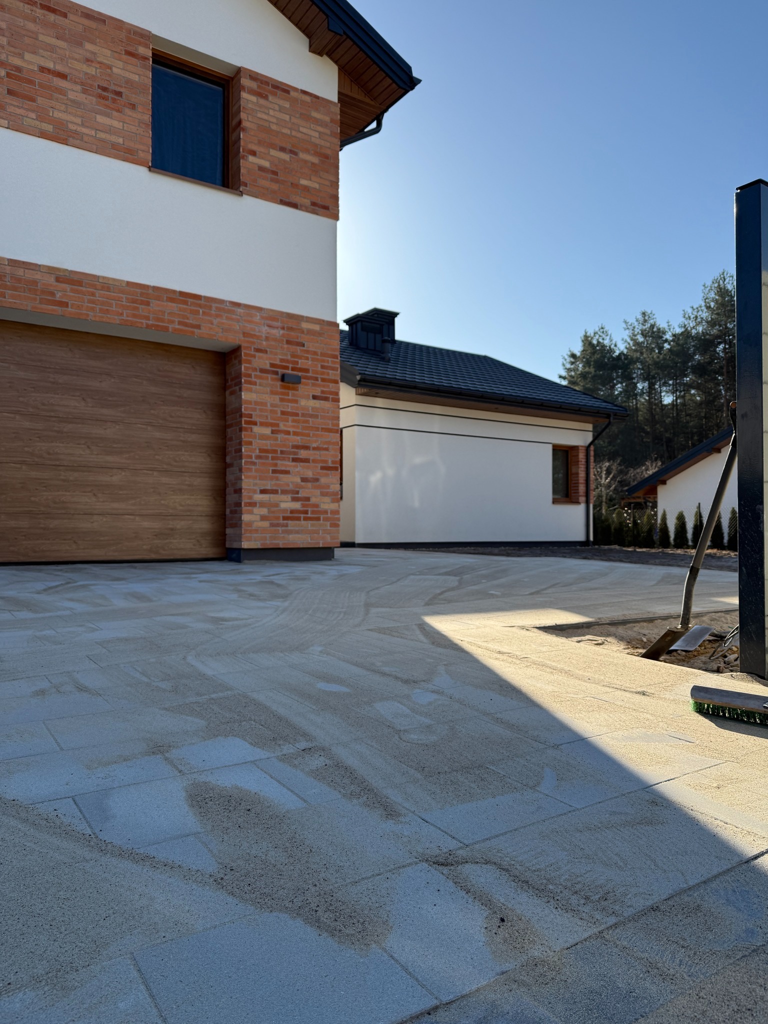 Nowoczesny dom z garażem i ułożoną kostką brukową, widoczne narzędzia brukarskie. Jasne, słoneczne oświetlenie podkreśla detale elewacji i wzór kostki.