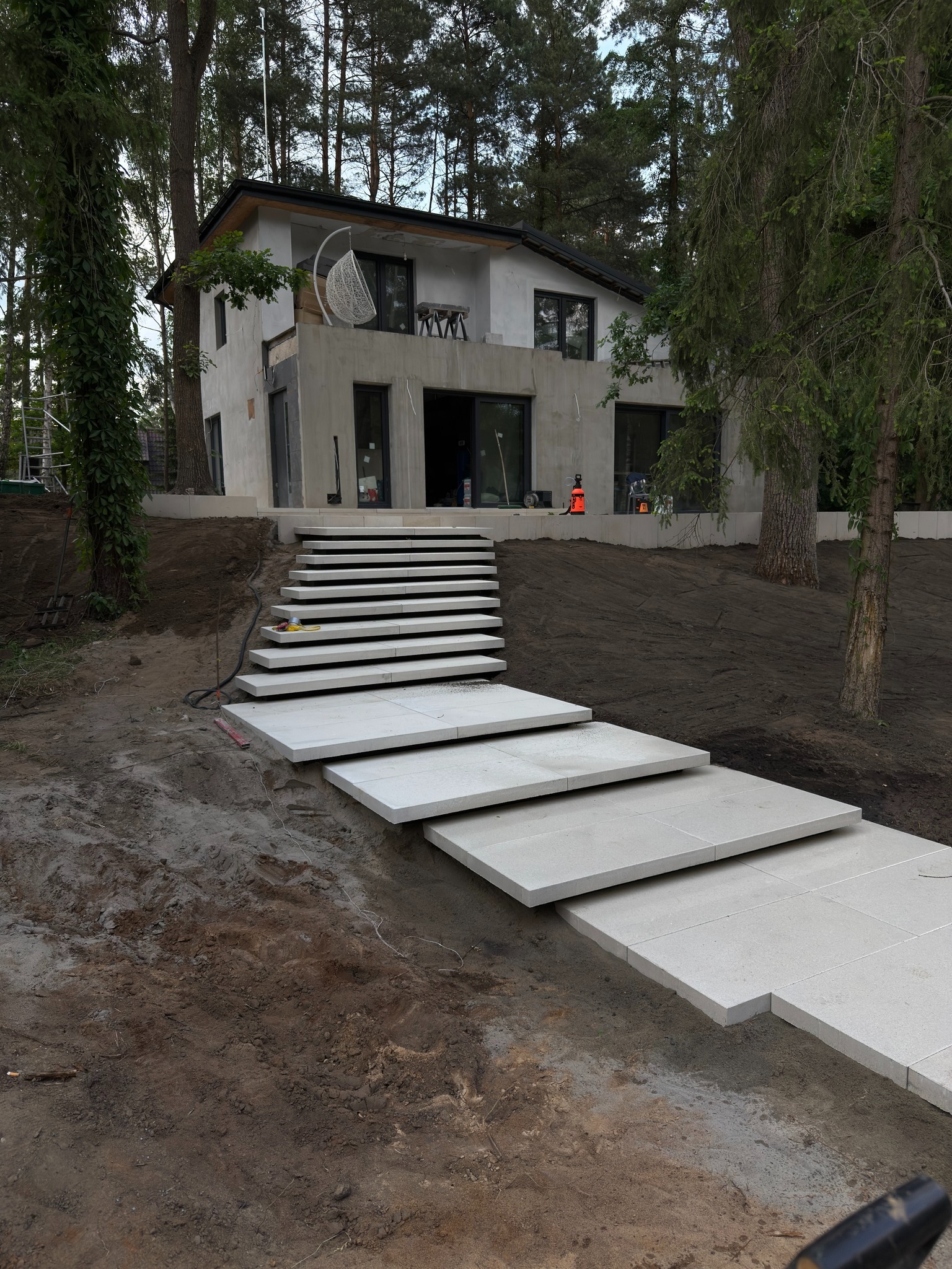 Nowoczesne schody z płyt kamiennych prowadzące do domu w otoczeniu drzew. Widoczne narzędzia i świeżo wyrównana ziemia.