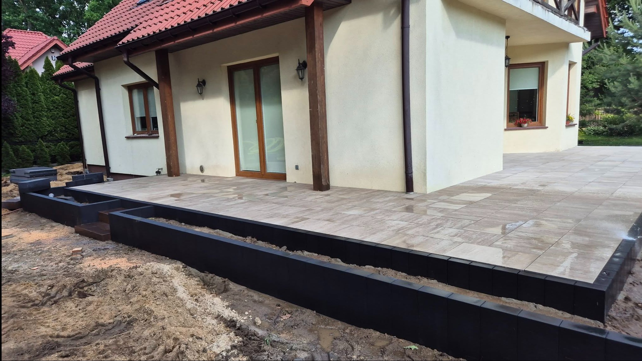 Jasna elewacja domu z tarasem wyłożonym kostką. Widoczne czarne obrzeża rabat i fragment ogrodu. Ujęcie z niskiej perspektywy podkreśla przestrzeń.