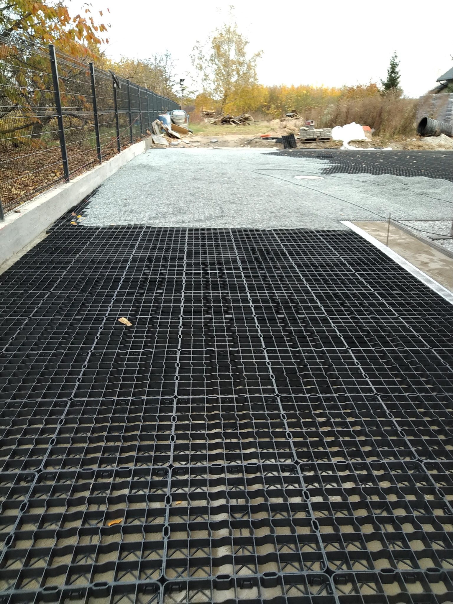 Geokrata na podsypce żwirowej, widok z góry. Fragment ogrodzenia w tle. Przygotowanie podłoża pod nawierzchnię przepuszczalną.
