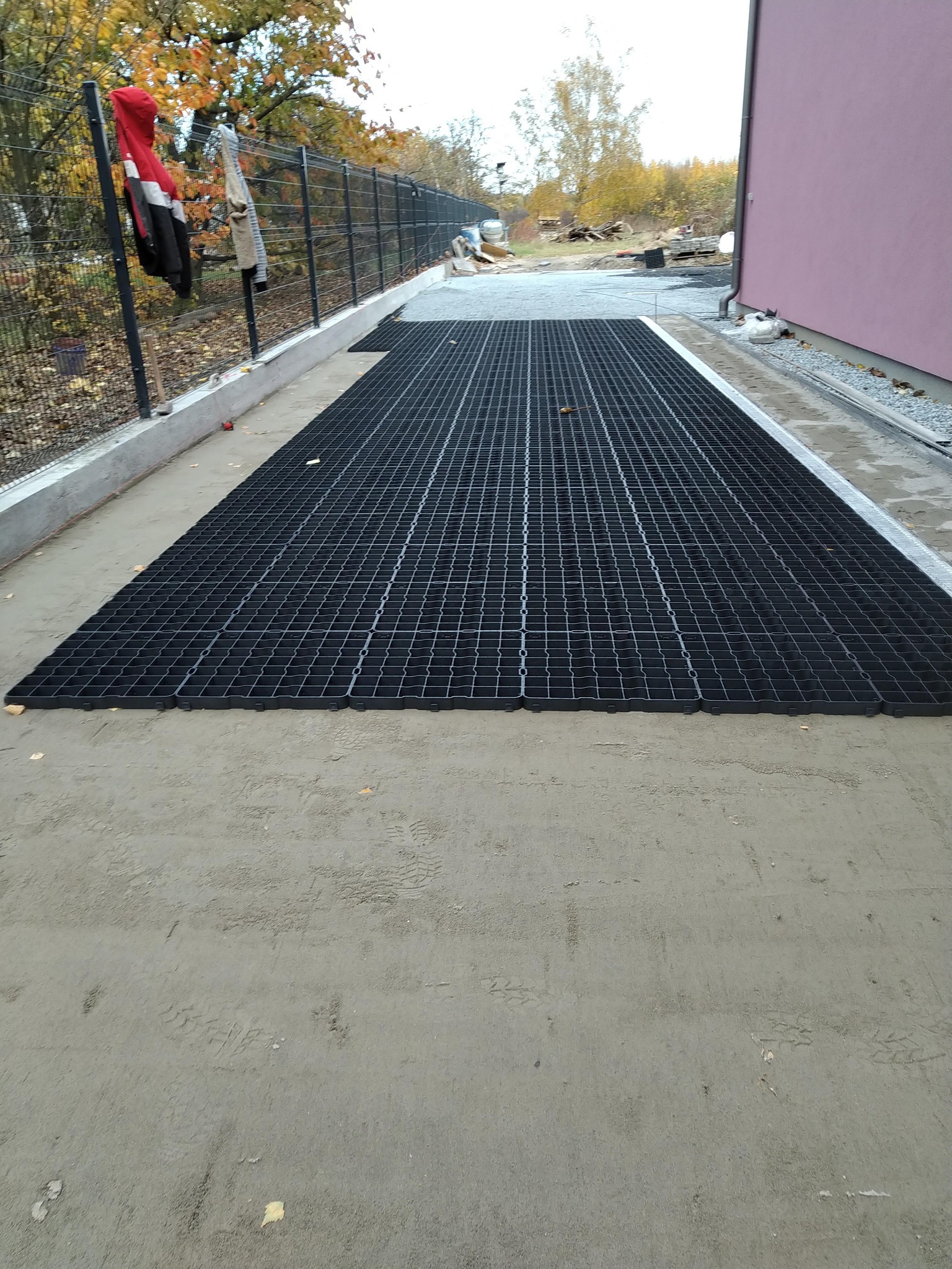 Ułożona, czarna kratka trawnikowa na przygotowanym podłożu z piasku. W tle ogrodzenie z siatki i budynek z różową elewacją. Widok z góry.