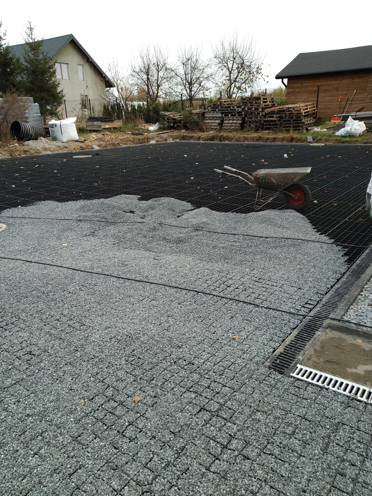 Wypełnianie czarnej kratownicy grysem. Widoczny fragment korytka odwadniającego, taczka i budynki w tle. Etap prac przygotowawczych podłoża.