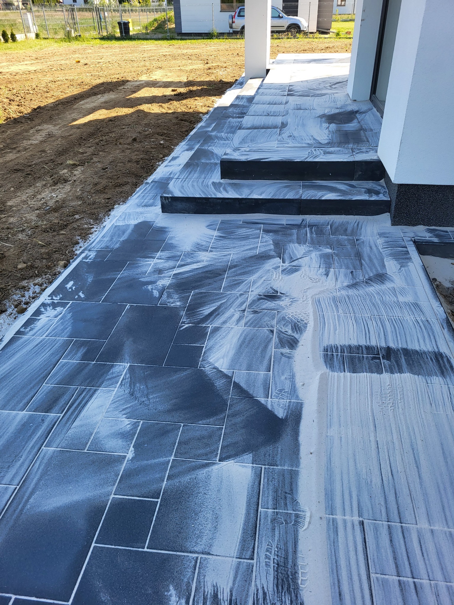 Świeżo ułożona, ciemna kostka brukowa przed wejściem do domu, jeszcze mokra po czyszczeniu. Widoczne schody z kostki i fragment trawnika. Jasne kolumny podtrzymują zadaszenie.