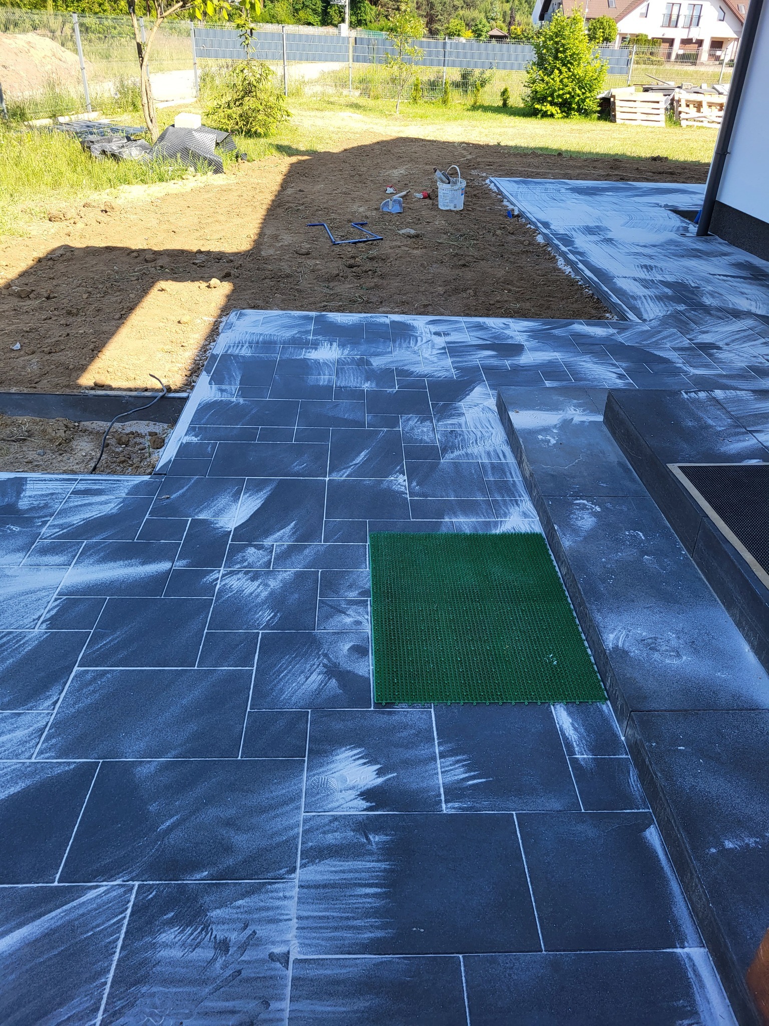 Częściowo ułożona, grafitowa kostka brukowa na posesji, z widocznym fragmentem trawnika i narzędziami. Etap prac z białym nalotem na powierzchni kostki.