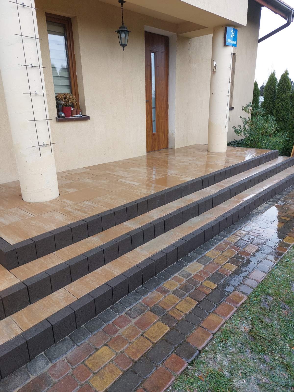 Ułożone schody wejściowe z kostki brukowej w odcieniach beżu i brązu, z ciemnymi obrzeżami. Widoczny fragment chodnika z kolorowej kostki. Słup z numerem domu.
