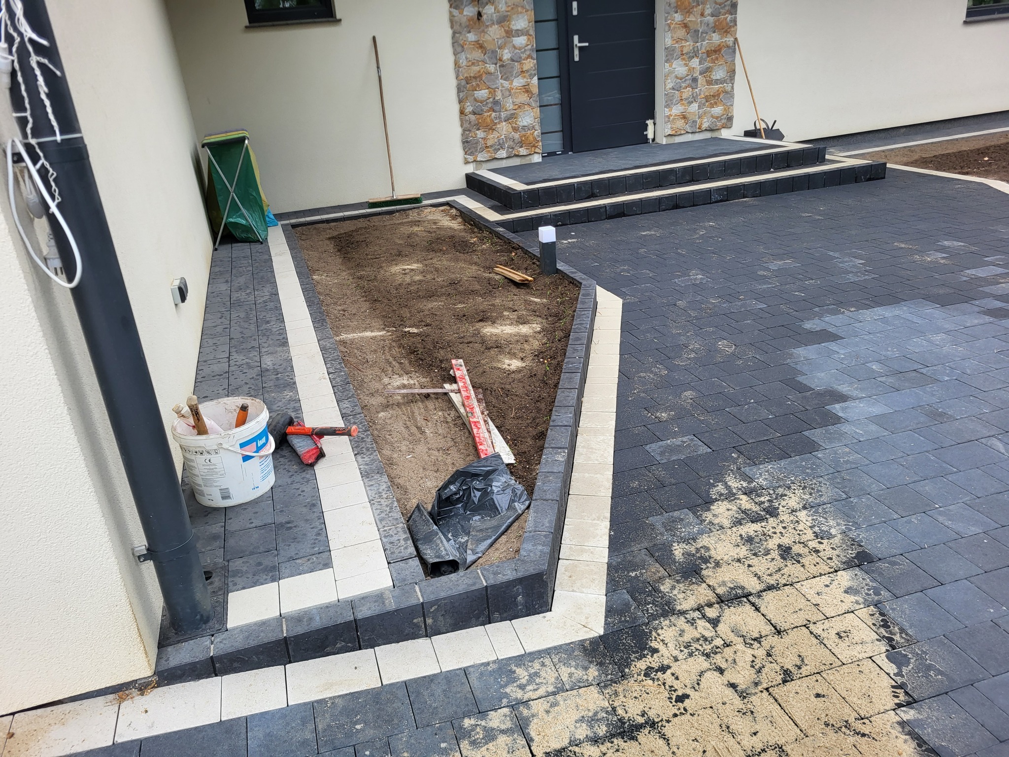 Częściowo ułożona, grafitowa kostka brukowa z jasnymi obrzeżami przed domem. Widoczny fragment rabaty, narzędzia i wiadro. Etap prac brukarskich.