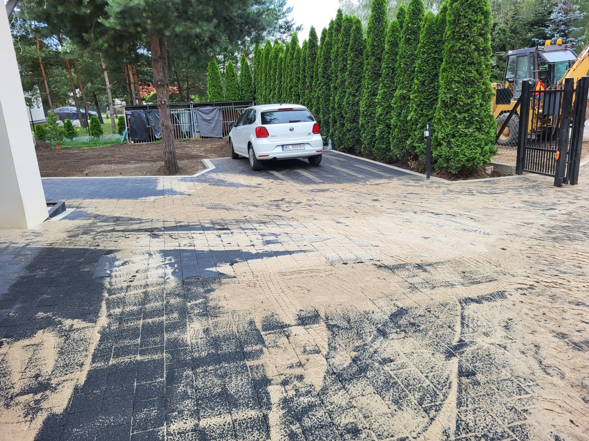 Ciemna kostka brukowa na podjeździe, częściowo zasypana piaskiem, biały samochód w tle, rząd tuj i brama wjazdowa. Widoczny sprzęt budowlany.