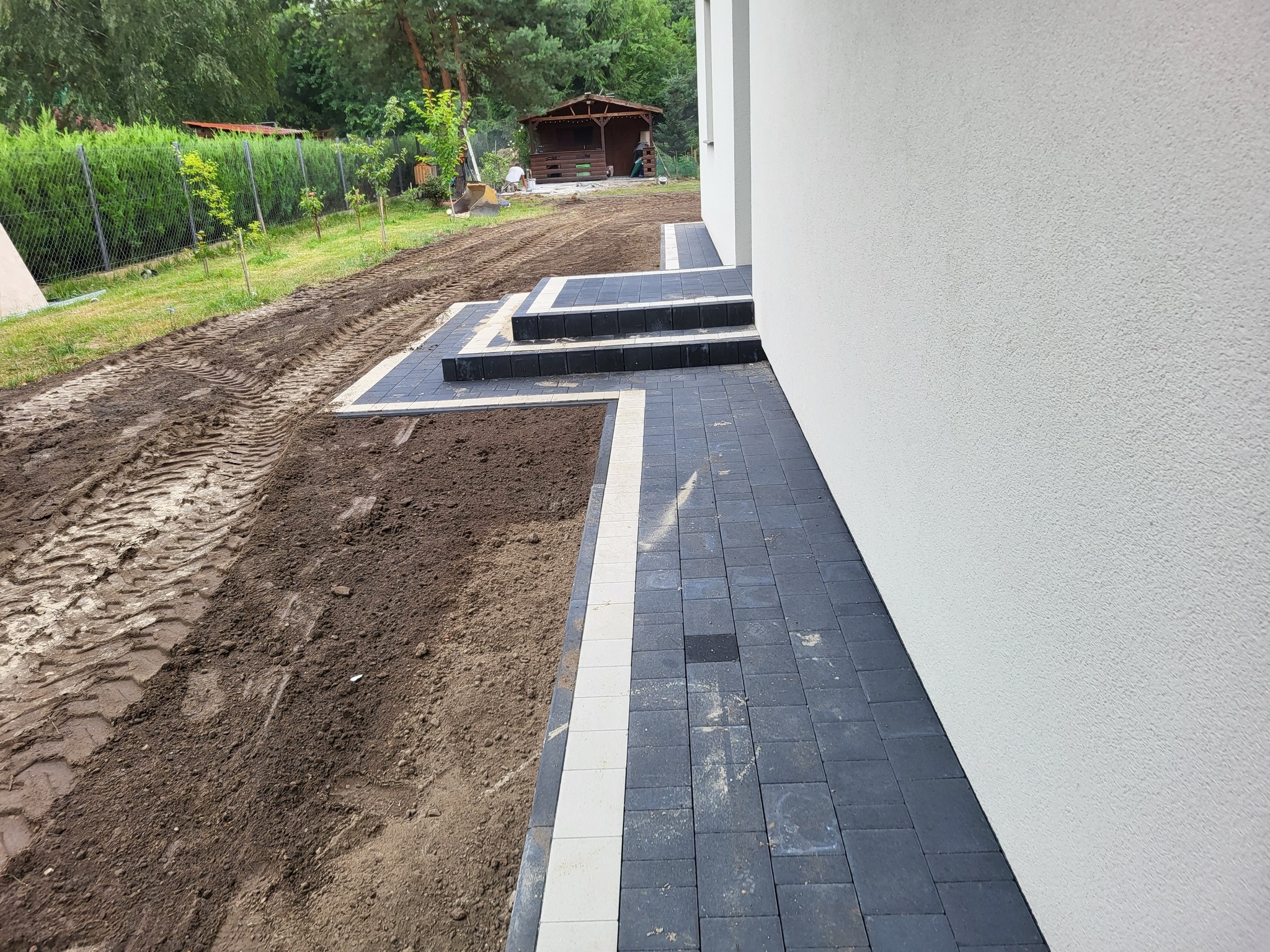 Nowoczesna ścieżka z kostki brukowej w odcieniach szarości i bieli, prowadząca do budynku z jasną elewacją. Widoczne schody i świeżo przygotowany teren pod trawnik.