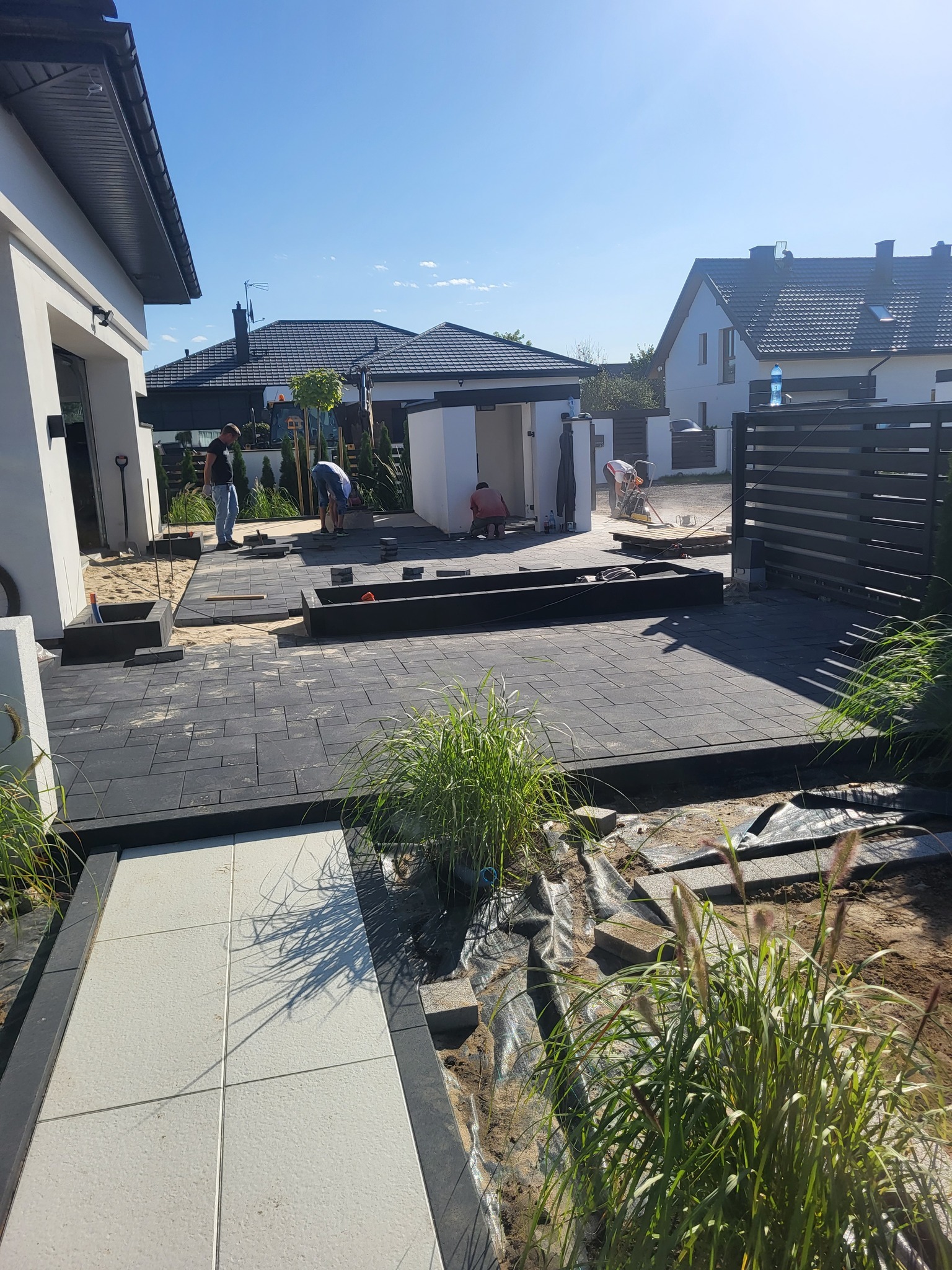 Układanie ciemnej kostki brukowej na posesji domu jednorodzinnego. Widoczne elementy ogrodu i pracujący robotnicy. Częściowo ułożona nawierzchnia.