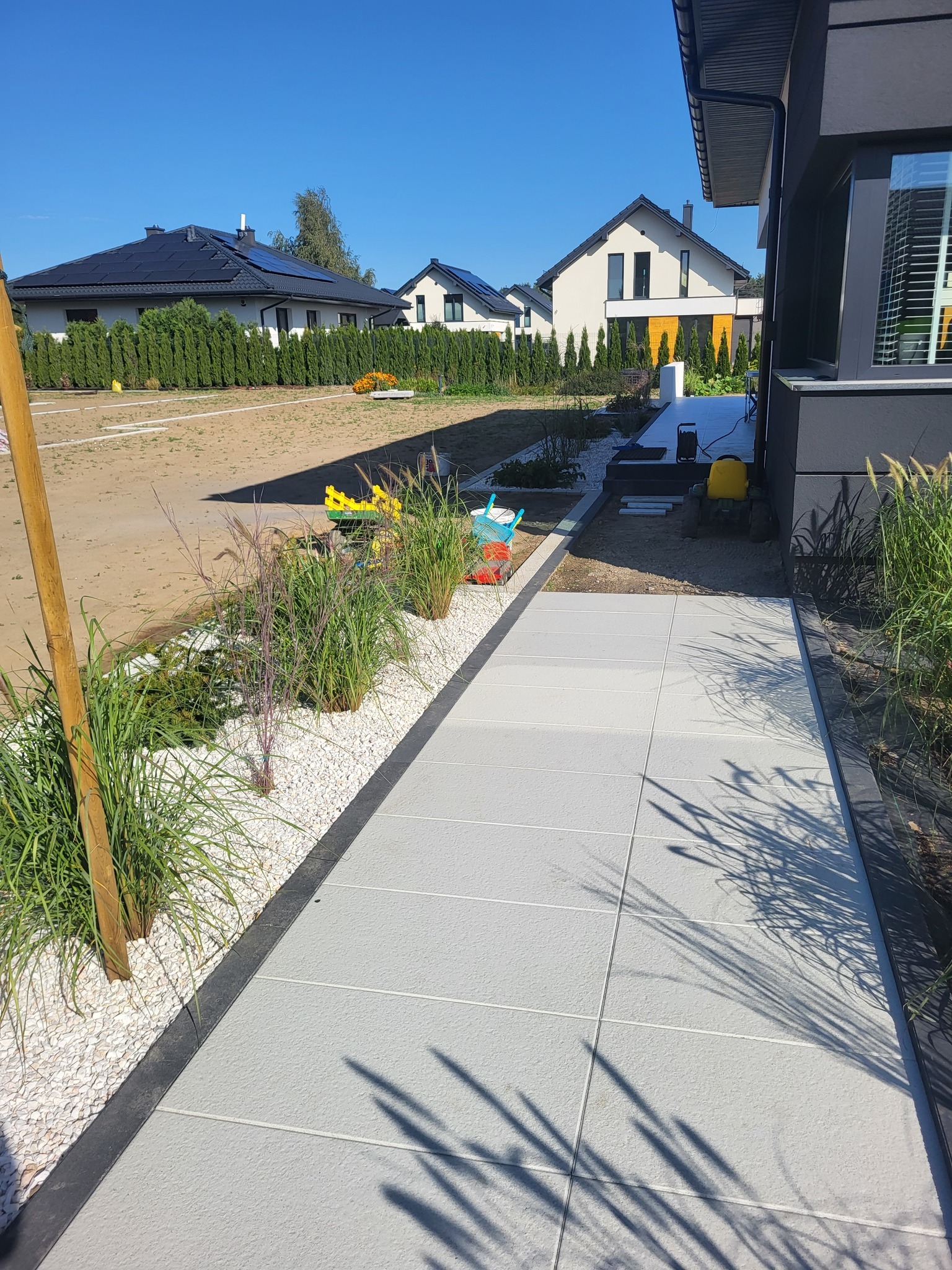 Nowoczesny chodnik z jasnych płyt, obrzeża z ciemnej kostki, biały żwir i ozdobne trawy. W tle domy jednorodzinne i błękitne niebo.