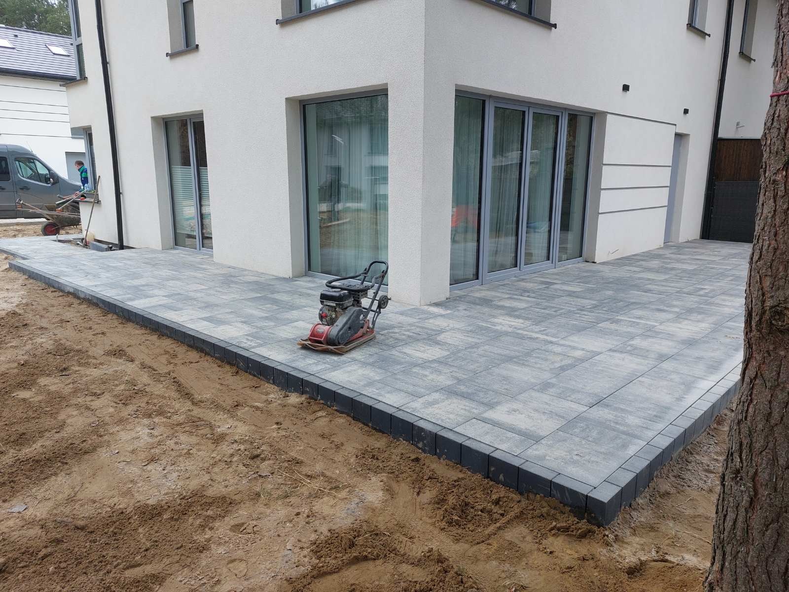 Szara kostka brukowa ułożona przy nowoczesnym domu z dużymi oknami. Na kostce stoi zagęszczarka. Widoczny fragment niezakończonej nawierzchni z ziemią.