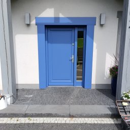 stanislaw werbowecki - Błękitne drzwi wejściowe z szybą, otoczone ramą w tym samym kolorze, na tle białej ściany z lampami. Widoczna kostka brukowa i narzędzia.