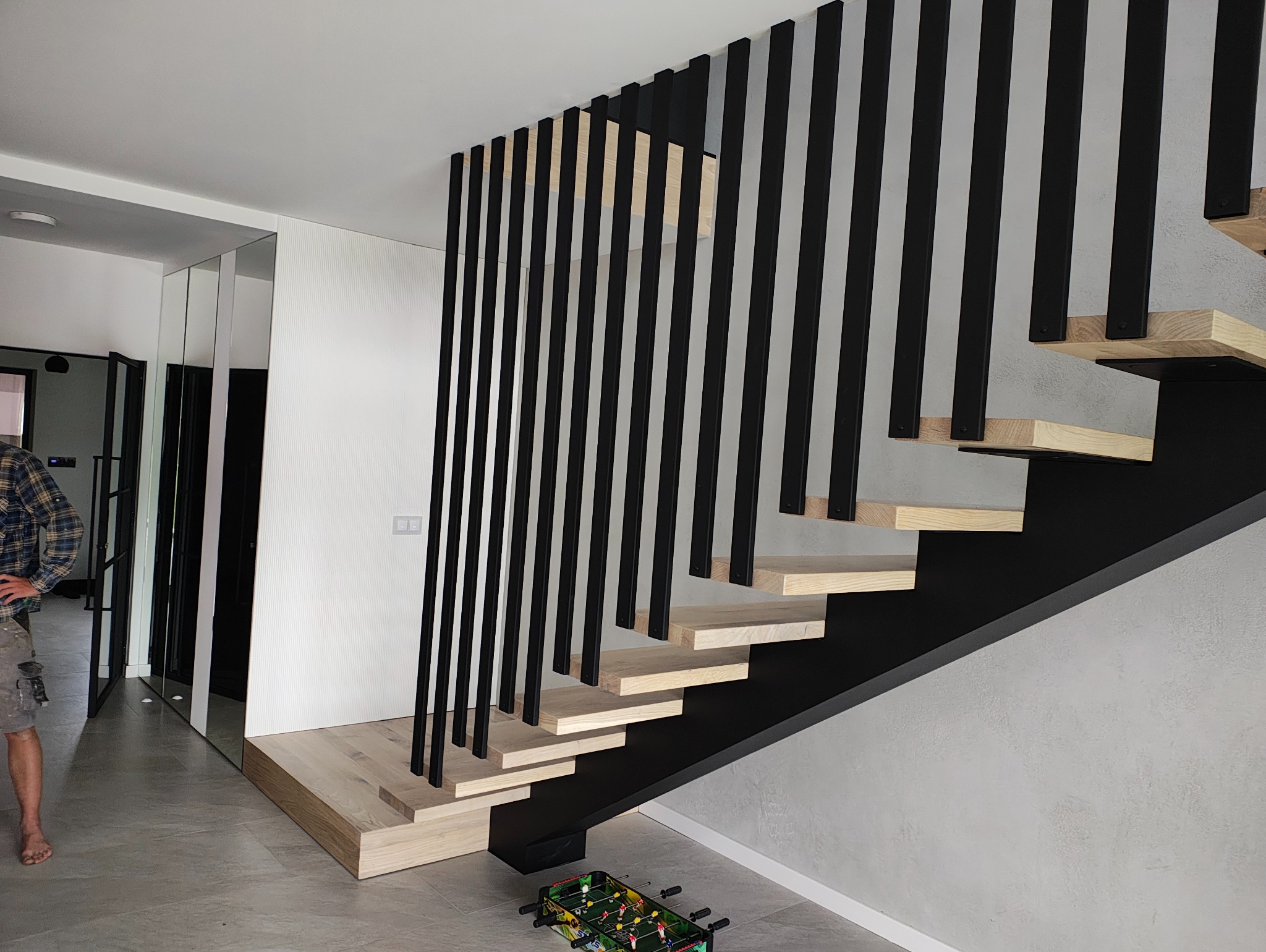 Nowoczesne schody z drewnianymi stopniami i czarną, pionową balustradą. Minimalistyczny design, jasne drewno kontrastuje z ciemną konstrukcją. W tle widoczne lustra.