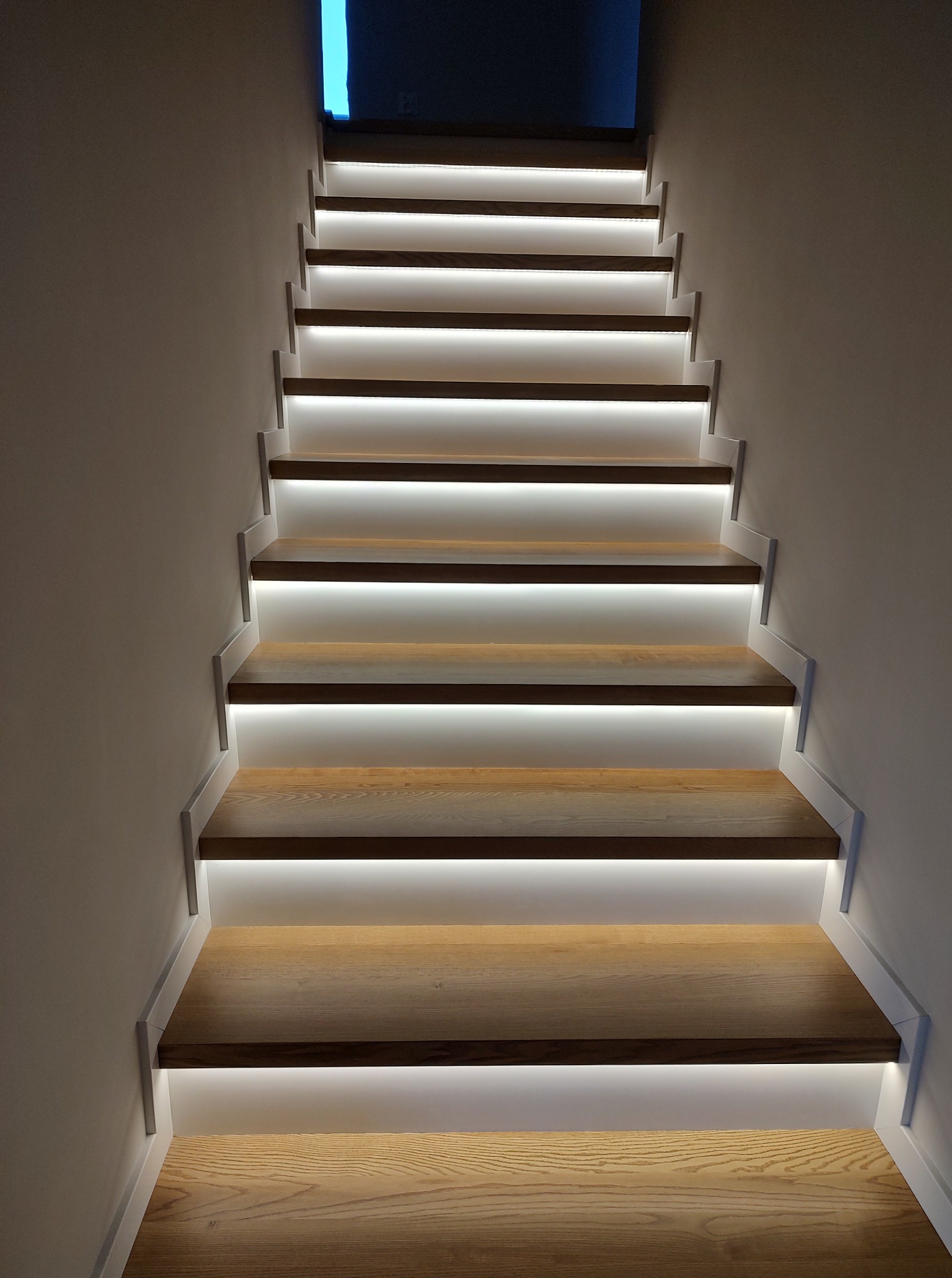 Nowoczesne schody drewniane z subtelnym podświetleniem LED, widok z dołu, podkreślający minimalistyczny design i ciepłe odcienie drewna. Oświetlenie akcentuje strukturę.