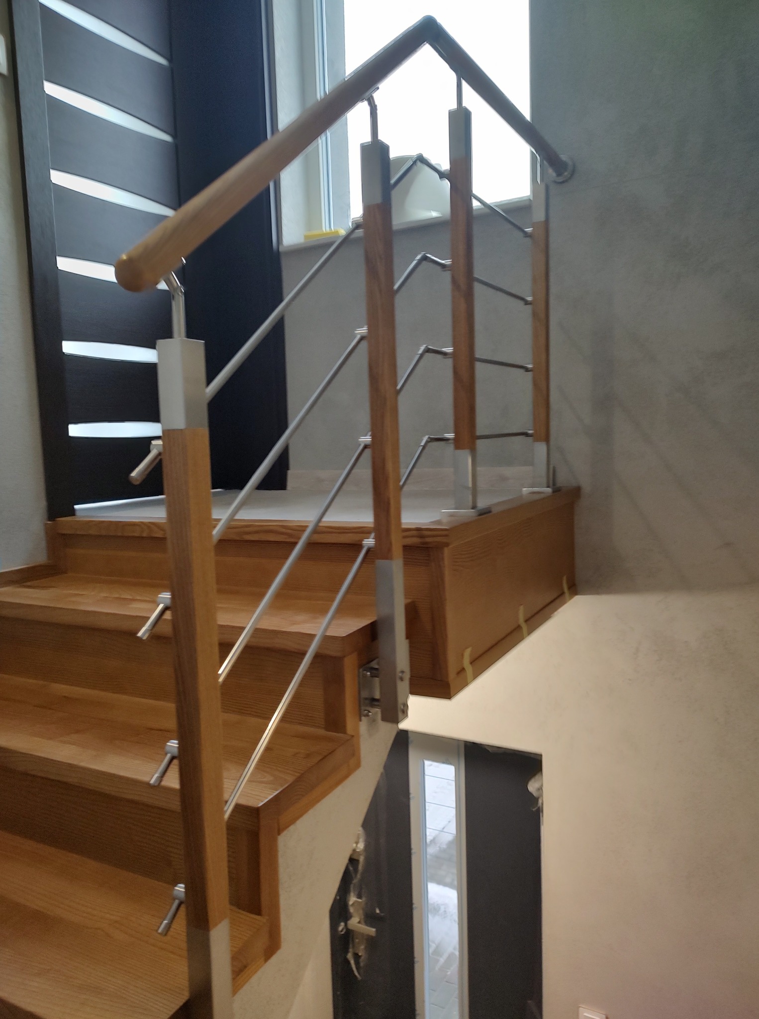 Jasne schody z drewnianymi stopniami i metalowymi poręczami, widoczne drzwi w tle. Ujęcie z dołu, podkreślające konstrukcję schodów i nowoczesny design.