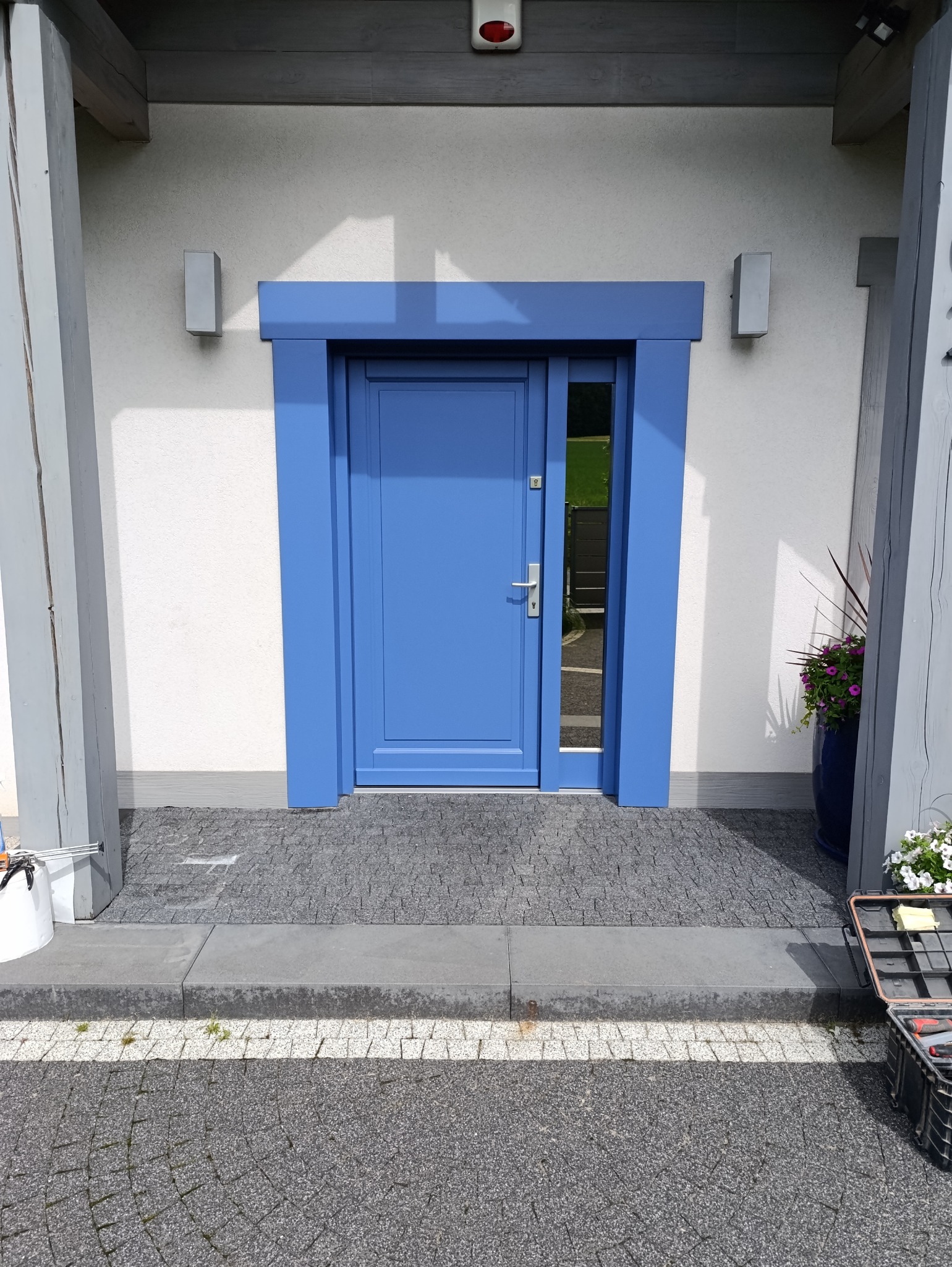 Błękitne drzwi wejściowe z szybą, otoczone ramą w tym samym kolorze, na tle białej ściany z lampami. Widoczna kostka brukowa i narzędzia.