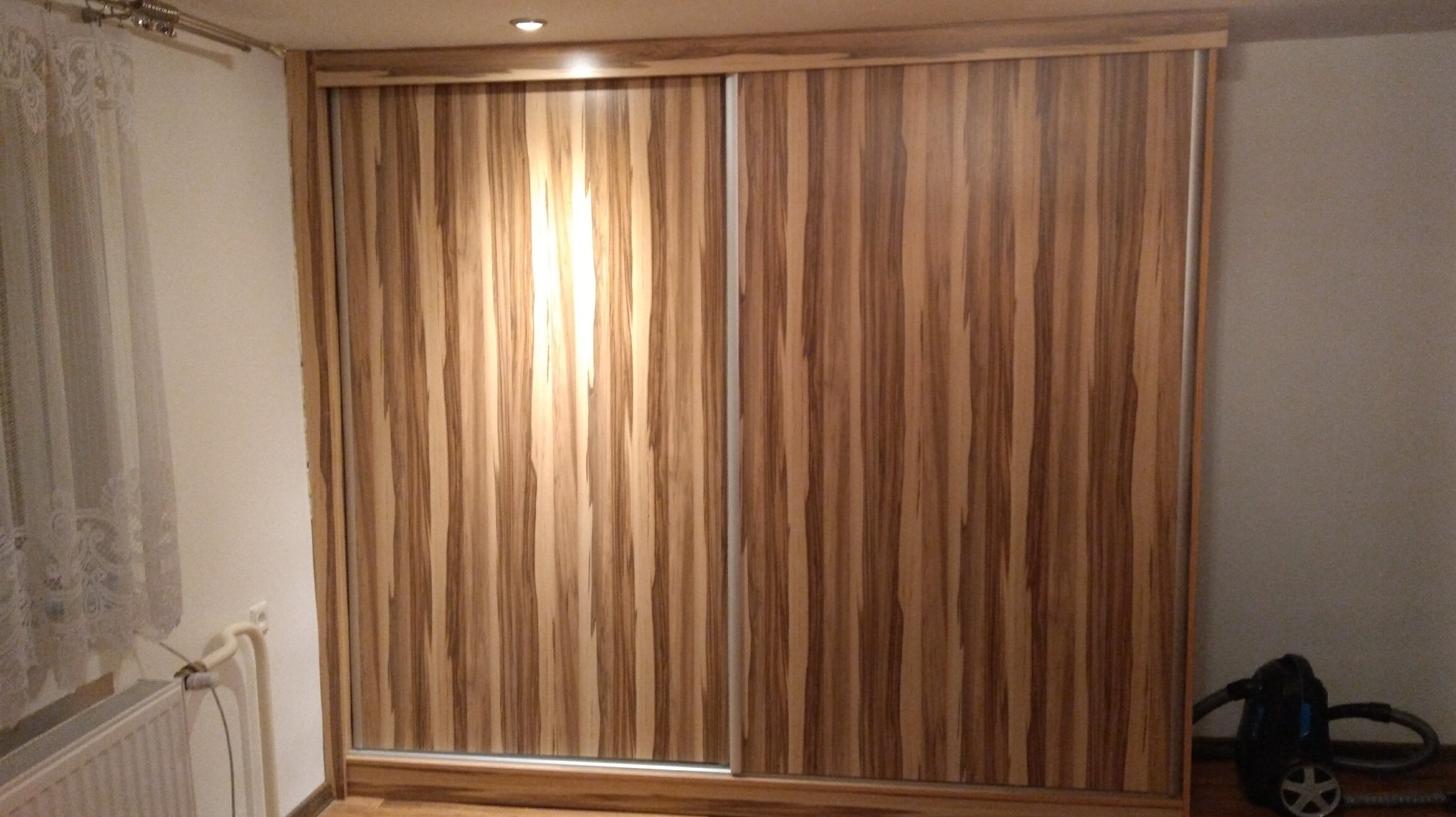 Szafa przesuwna z drewnopodobnymi frontami, widoczne pionowe pasy w odcieniach brązu i beżu, srebrne prowadnice, na podłodze odkurzacz.