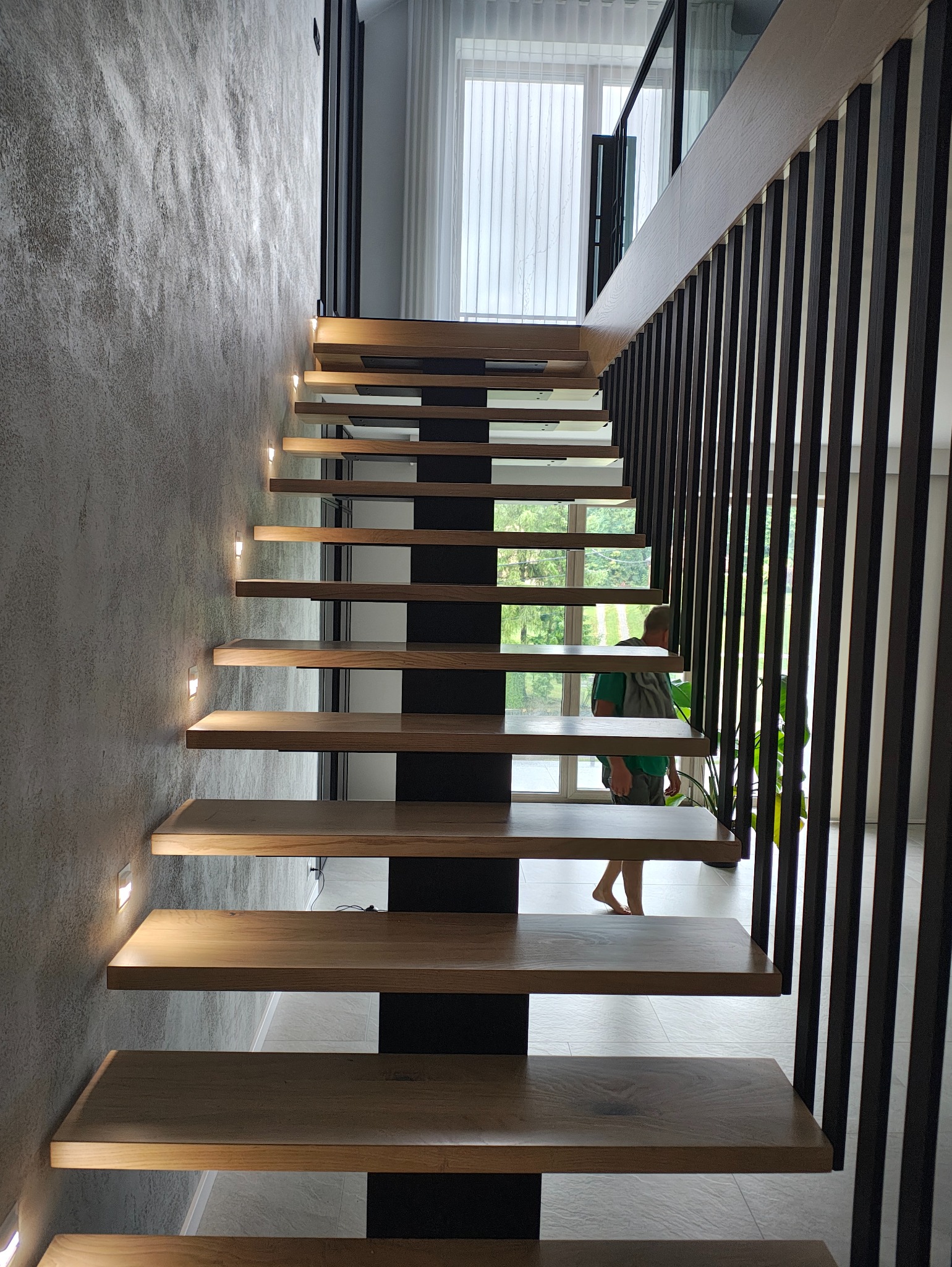 Nowoczesne schody z drewnianymi stopniami i podświetleniem LED na ścianie o fakturze betonu. Minimalistyczna balustrada z pionowych, czarnych elementów.