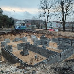 Biotech - Wykopane fundamenty z częściowo wymurowanymi ścianami z szarych bloczków betonowych. Na drugim planie zabudowa wiejska i firmowy bus.