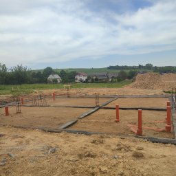 Biotech - Fundamenty domu jednorodzinnego w trakcie budowy, widoczne zbrojenia i instalacje kanalizacyjne, w tle krajobraz wiejski z zabudowaniami.