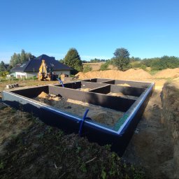 Biotech - Zalane fundamenty domu jednorodzinnego z hydroizolacją, widoczne niebieskie rury, w tle koparka i dom w budowie, krajobraz wiejski, słoneczny dzień.