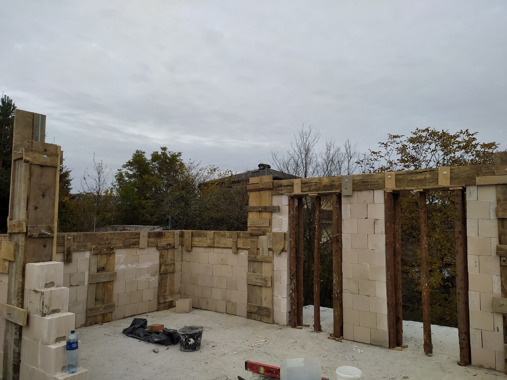 Wznoszenie ścian z betonu komórkowego Ytong. Widoczne drewniane szalunki i podpory. Na podłożu narzędzia murarskie i butelka wody. Prace murarskie w toku.