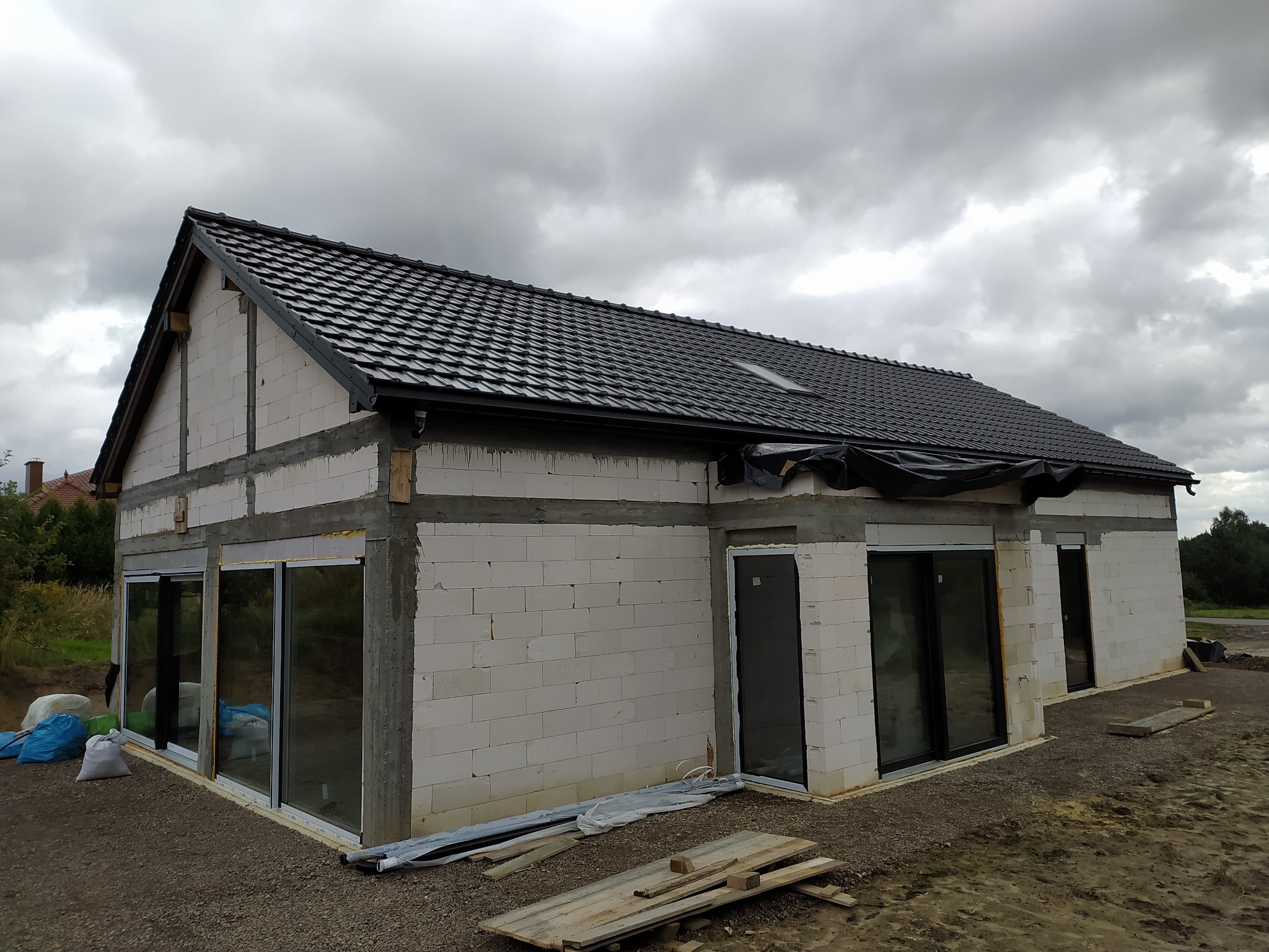Niedokończony dom z białych bloczków, czarnym dachem i dużymi oknami przesuwnymi. Widoczne drewniane elementy konstrukcyjne i gruz budowlany. Pochmurne niebo w tle.