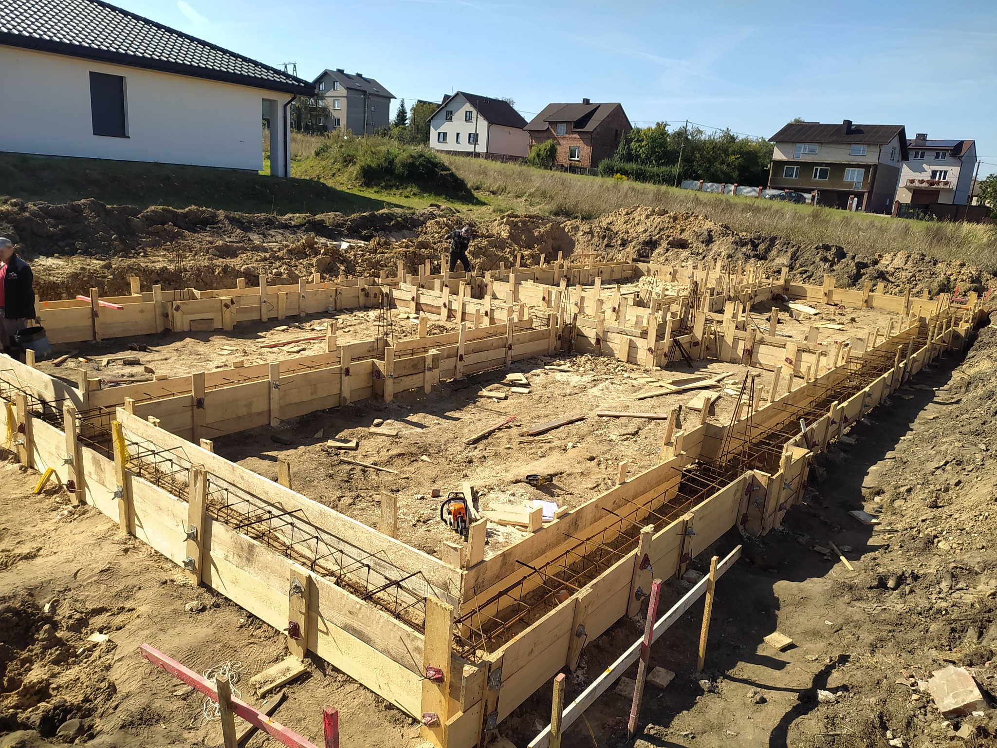Zalane fundamenty domu jednorodzinnego w drewnianym szalunku z widocznym zbrojeniem, na tle zabudowy mieszkalnej i błękitnego nieba.