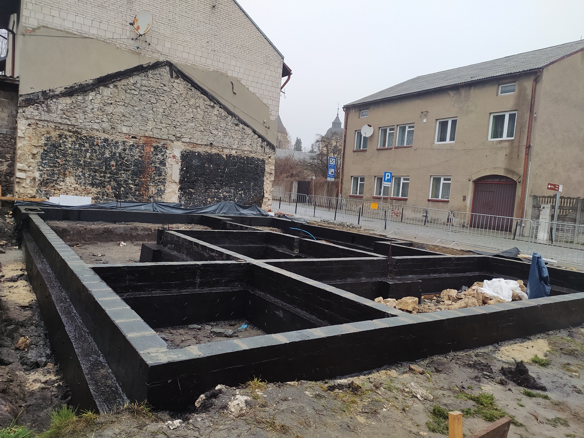Zabezpieczone fundamenty budynku w trakcie budowy, pomalowane na czarno, widoczne otoczenie placu budowy i budynki w tle. Ujęcie z poziomu gruntu.