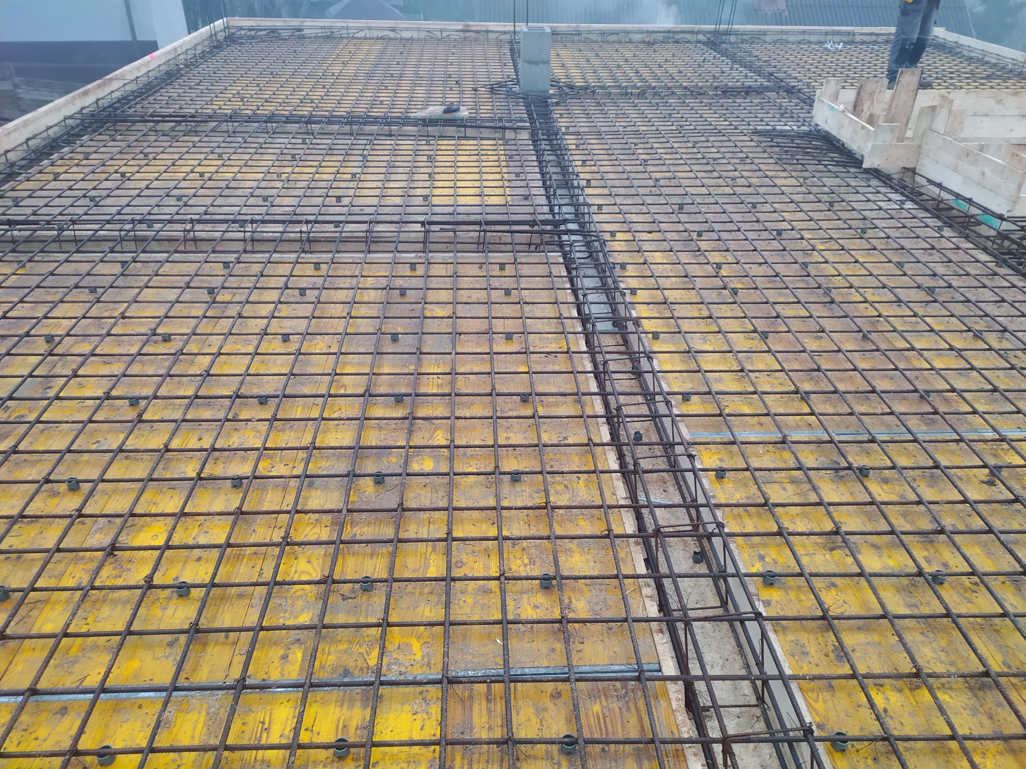 Zbrojenie stropu z żółtymi deskami szalunkowymi, widok z góry. Stalowe pręty tworzą siatkę, betonowe elementy konstrukcyjne w tle. Etap budowy.