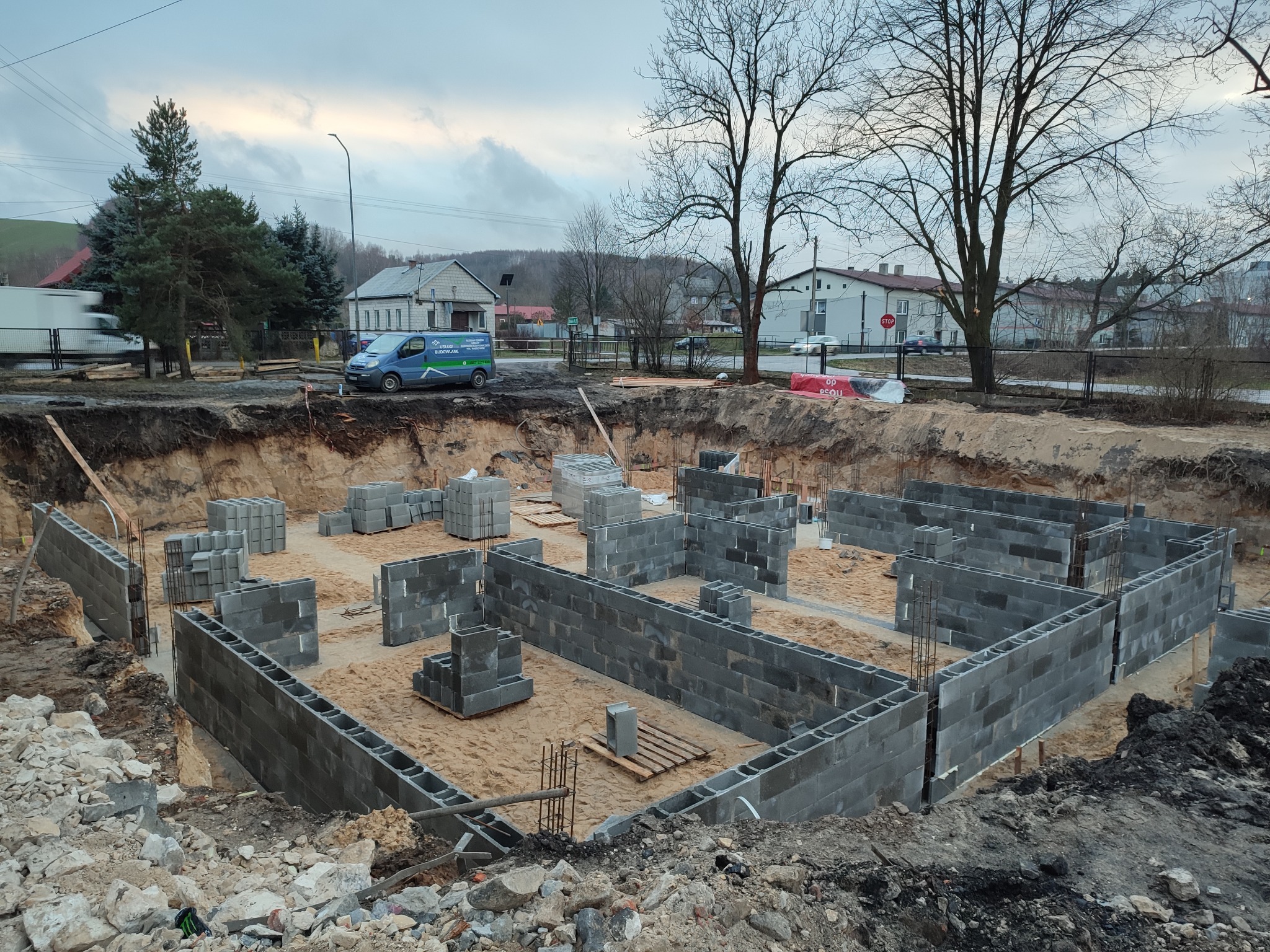 Wykopane fundamenty z częściowo wymurowanymi ścianami z szarych bloczków betonowych. Na drugim planie zabudowa wiejska i firmowy bus.