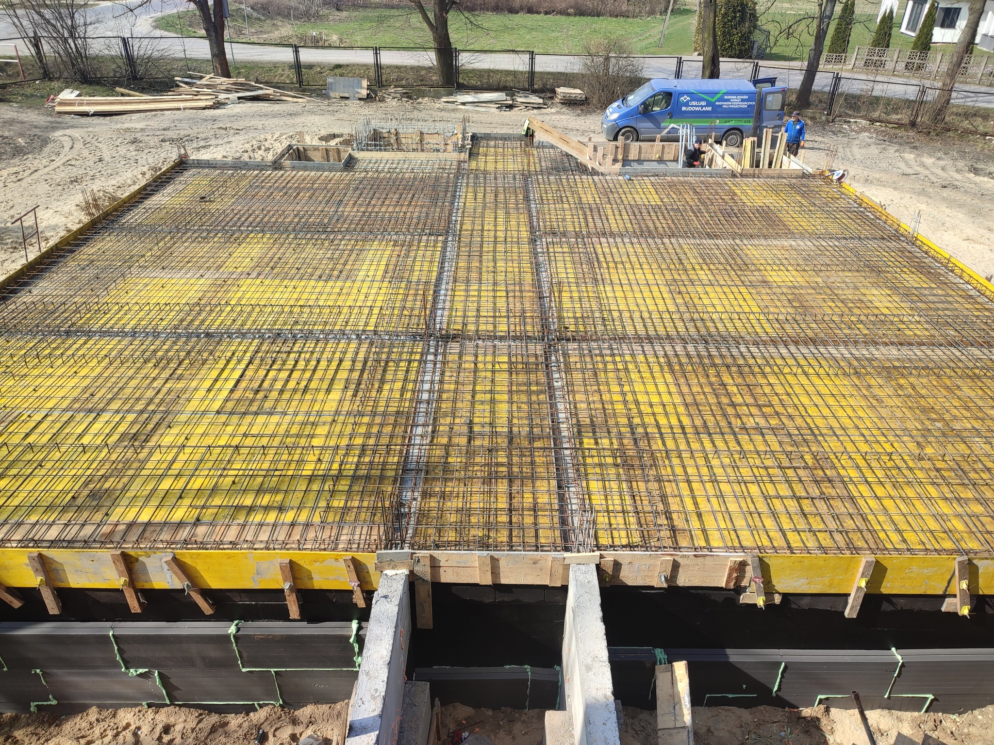 Zbrojenie fundamentów z żółtym szalunkiem i stalową siatką, przygotowane do wylania betonu. W tle widoczny samochód budowlany i dwóch pracowników.