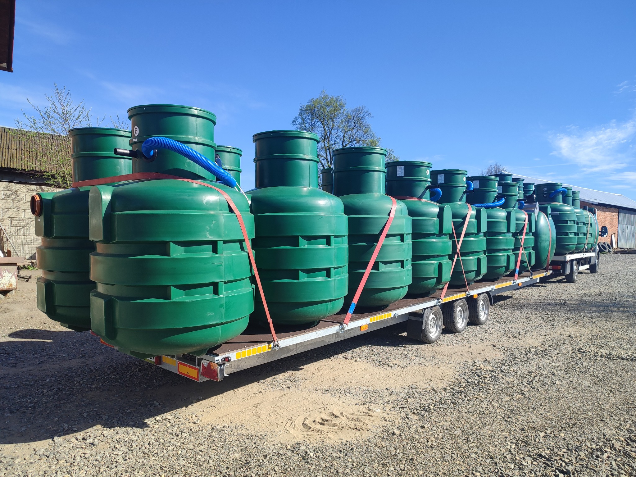 Transport zbiorników na szambo na przyczepie, zabezpieczonych pasami. Zielone zbiorniki z niebieskimi rurami na tle błękitnego nieba i budynków.